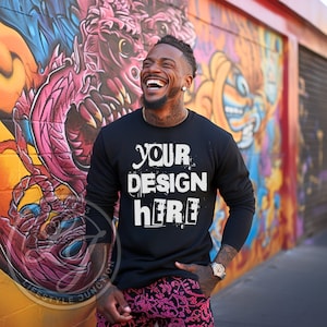 Pode incluir: Uma camisa de manga comprida preta com o texto "YOUR DESIGN HERE" em branco, letras desgastadas. A camisa está sendo usada por uma pessoa em pé na frente de uma parede com grafites coloridos.