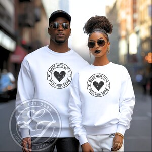 May include: A white sweatshirt with a black circle design featuring a heart and the text "Made with Love" repeated in a circular pattern. The text "Est. 2023" is visible below the design.