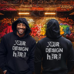 May include: Two black hooded sweatshirts with the text "YOUR DESIGN HERE" printed on the front and back. The sweatshirts are shown against a graffiti-covered wall.