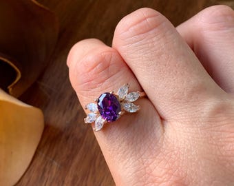 May include: A gold-toned ring featuring a central oval-cut purple gemstone flanked by three marquise-cut clear stones on each side. The band is thin and metallic, showcasing a vibrant violet center stone contrasted by the clear, faceted side accents.