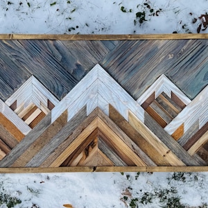 May include: Wooden mountain wall art with a framed design. The artwork features a mountain range constructed from various shades of wood, including gray, white, and brown. The mountains are set against a darker, gray-blue wooden background.
