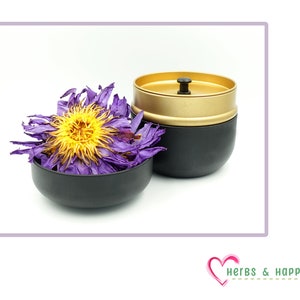 Puede incluir: Un recipiente negro y dorado con tapa y asa negras, junto a un peque&ntilde;o cuenco negro lleno de flores moradas y amarillas secas. La imagen est&aacute; sobre un fondo blanco.