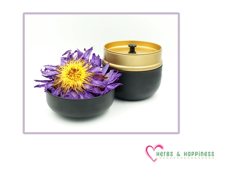 Puede incluir: Un recipiente negro y dorado con tapa y asa negras, junto a un peque&ntilde;o cuenco negro lleno de flores moradas y amarillas secas. La imagen est&aacute; sobre un fondo blanco.
