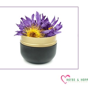 Puede incluir: Un recipiente negro y dorado lleno de una flor morada seca con un centro amarillo. El recipiente tiene una base negra y un borde dorado. La imagen est&aacute; sobre un fondo blanco. El logotipo de Herbs & Happiness est&aacute; en la esquina inferior derecha.
