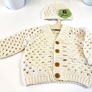 Puede incluir: Un cárdigan de punto blanco con cierre de botones y un gorro de punto blanco a juego con un adorno de flor verde.