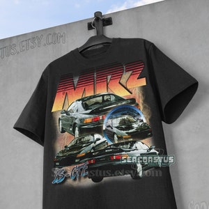 Puede incluir: Camiseta negra con un gráfico retro de tres coches y el texto "MR2" en una fuente audaz. El diseño incorpora un degradado de naranja, rojo y amarillo. El texto "3S-GTE" también es visible. La camiseta está colgada contra una pared gris.