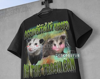 Könnte beinhalten: Schwarzes T-Shirt mit einer Grafik von zwei Opossums mit geröteten Wangen und dem Text "What If We Accidentally Kissed In The Trash Can" in Grün und Weiß. Das T-Shirt hängt vor einer grauen Wand.