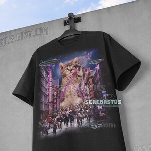 May include: Black t-shirt with a graphic print of a cat with laser beams shooting from its eyes. The cat is sitting in a city street with people running away from it. The text "Seacoastus" is printed on the bottom right corner of the shirt.