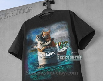 May include: Black t-shirt featuring a graphic of two cats embracing in a bucket labeled "Titanic." The image includes a bucket of fish and the text "SEACOASTUS.ETSY.COM."