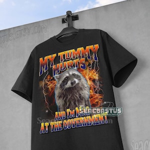 May include: Black t-shirt with a graphic of a raccoon with flames and lightning bolts. The text "My Tummy Hurts And I'm Mad At The Government" is printed on the shirt.