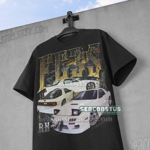 Op de afbeelding: Zwart T-shirt met een grafische print van witte sportwagens en de tekst "RX-7" in gouden letters. De tekst "SEACOASTUS" is ook zichtbaar op het T-shirt.