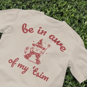 May include: A beige t-shirt with a red graphic of a cat wearing a wizard hat and holding a wand. The text "be in awe of my 'tsim" is printed on the shirt.
