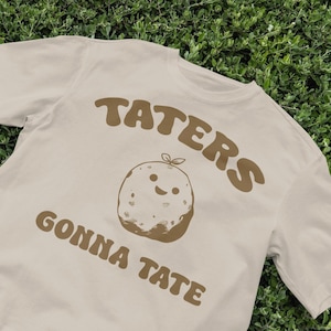 May include: A beige t-shirt with a brown graphic of a potato with a smiling face and the text "Taters Gonna Tate".