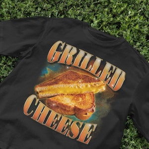 Può includere: Maglietta nera con una grafica di un panino al formaggio grigliato e il testo "GRILLED CHEESE" in stile retrò.