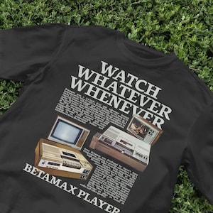 May include: Black t-shirt featuring a vintage advertisement for a Betamax player. The ad shows a black and white television and a Betamax player with the text "Watch Whatever Whenever" and "Betamax Player".