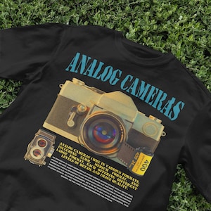May include: Black t-shirt with a graphic of an analog camera and film roll. The text "ANALOG CAMERAS" is printed in blue on the shirt. The text below the image reads "ANALOG CAMERAS COME IN VARIOUS FORMATS, INCLUDING 35MM, MEDIUM FORMAT, AND LARGE FORMAT, EACH OFFERING DIFFERENT LEVELS OF DETAIL AND IMAGE QUALITY."