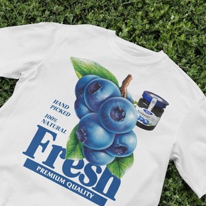 Op de afbeelding: Wit T-shirt met een afbeelding van blauwe bessen en een potje blauwe bessenjam. De tekst "Fresh Premium Quality" staat op het shirt gedrukt. De tekst "Hand Picked 100% Natural" staat ook op het shirt gedrukt.