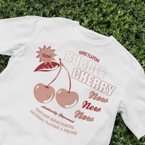 Könnte beinhalten: Weißes T-Shirt mit einer roten und weißen Grafik von zwei Kirschen und dem Text "SUPER FLAVOUR BUBBLE CHERRY New New New Artilically Flavoured CONTAINS SUGAR GLUCOSE, ARTIFICAL FLAVOUR & COLOUR."