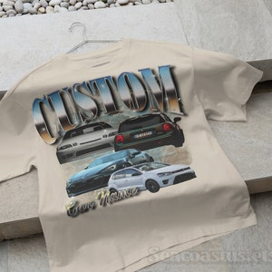 以下が含まれることがあります： ベージュのTシャツに、4台の車が描かれたグラフィックプリントと、スタイリッシュなフォントで「CUSTOM」という文字がプリントされています。「Car Mania」という文字が車の下にプリントされています。