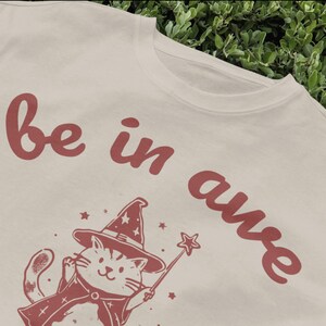 May include: A white t-shirt with a red graphic of a cat wearing a witch hat and holding a wand. The text "be in awe" is written in red on the shirt.