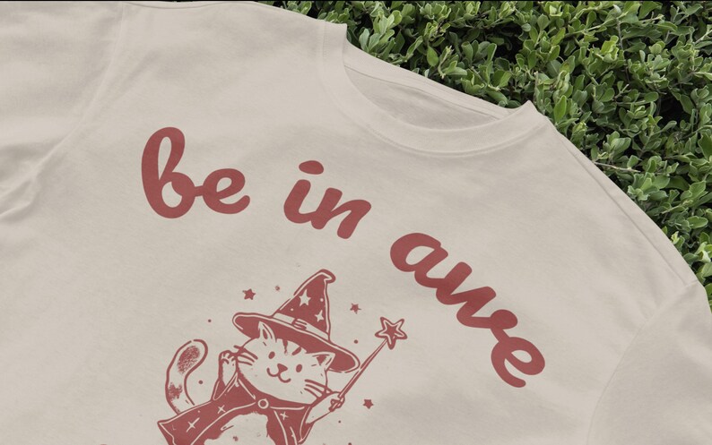 May include: A white t-shirt with a red graphic of a cat wearing a witch hat and holding a wand. The text "be in awe" is written in red on the shirt.