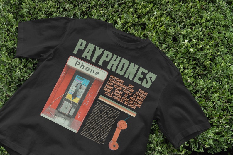 May include: Black t-shirt featuring a graphic of a payphone with the word "PAYPHONES" in green block letters. The shirt includes text explaining what payphones are and how they work.
