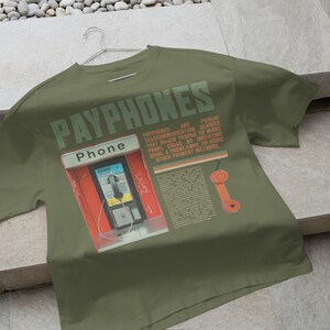 May include: Olive green t-shirt featuring a graphic of a red payphone booth with the word "Phone" above it. The shirt has the word "PAYPHONES" printed across the top in green, along with text and an orange phone receiver graphic.