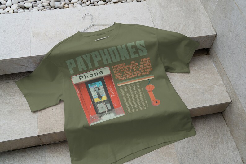 May include: Olive green t-shirt featuring a graphic of a red payphone booth with the word "Phone" above it. The shirt has the word "PAYPHONES" printed across the top in green, along with text and an orange phone receiver graphic.
