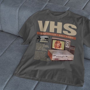 May include: A dark gray t-shirt with a vintage advertisement for a VHS video cassette recorder. The ad features a woman smiling in front of a television and the text "VHS Triple-System/Drei-Norm".
