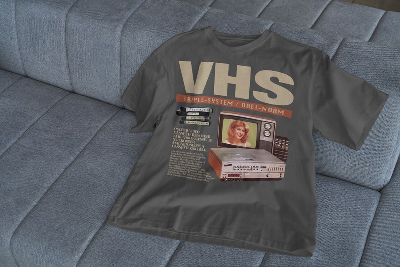 May include: A dark gray t-shirt with a vintage advertisement for a VHS video cassette recorder. The ad features a woman smiling in front of a television and the text "VHS Triple-System/Drei-Norm".