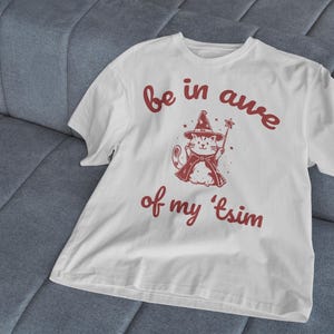 May include: White t-shirt with a red graphic of a cat wearing a wizard hat and holding a wand. The text "be in awe of my 'tsim" is printed in red below the cat.
