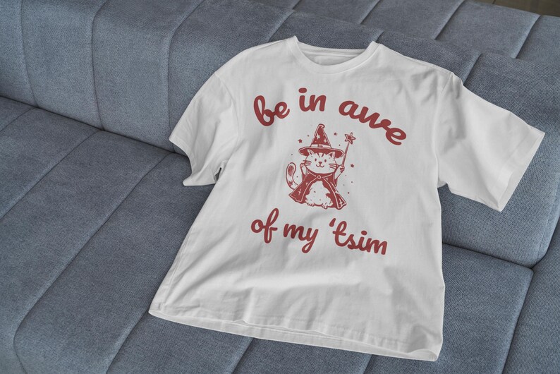 May include: White t-shirt with a red graphic of a cat wearing a wizard hat and holding a wand. The text "be in awe of my 'tsim" is printed in red below the cat.