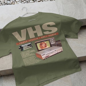 May include: A green t-shirt with a vintage VHS video cassette recorder and television set graphic. The text "VHS Triple-System / Orei-Norm" is printed on the shirt.