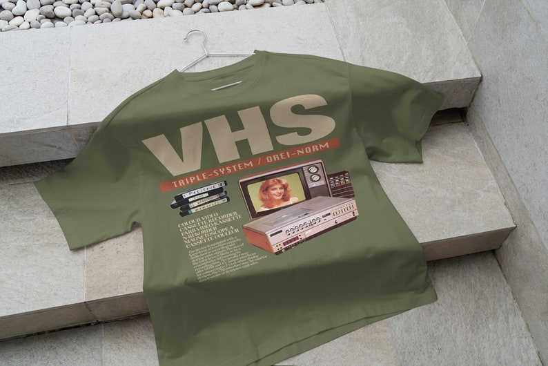 May include: A green t-shirt with a vintage VHS video cassette recorder and television set graphic. The text "VHS Triple-System / Orei-Norm" is printed on the shirt.