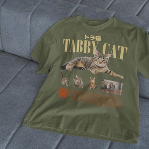 May include: A green t-shirt with a graphic print of tabby cats. The text "Tabby Cat" is printed in gold letters with the Japanese word for tabby cat above it.