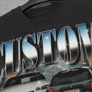 以下が含まれることがあります： 黒いTシャツに、銀色と青色の「CUSTOM」のグラフィックが施されています。グラフィックには、白いセダンと濃い緑色のハッチバックの2台の車が描かれています。グラフィックには、稲妻のデザインが施されています。