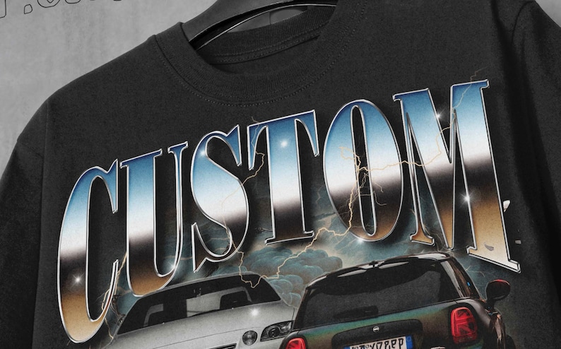 以下が含まれることがあります： 黒いTシャツに、銀色と青色の「CUSTOM」のグラフィックが施されています。グラフィックには、白いセダンと濃い緑色のハッチバックの2台の車が描かれています。グラフィックには、稲妻のデザインが施されています。