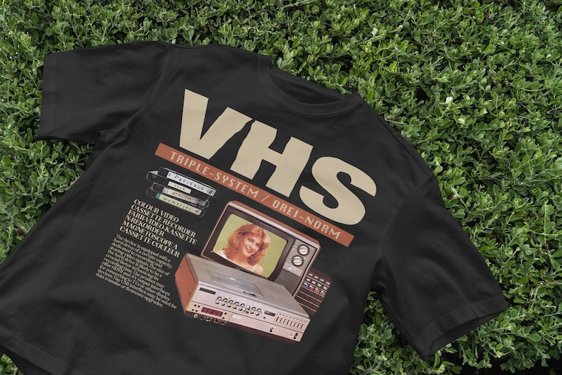 May include: Black t-shirt with a vintage VHS video cassette recorder advertisement. The text reads "VHS Triple-System/OREI-NORM" and features a color video cassette recorder, a television, and a cassette.
