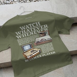 May include: A green t-shirt featuring a vintage advertisement for a Betamax player. The text reads "Watch Whatever Whenever Betamax Player".