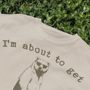 May include: A beige t-shirt with a brown bear wearing sunglasses and the text "I'm about to get" printed on it.