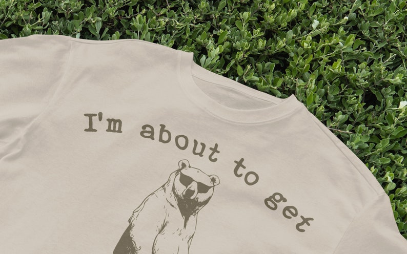 May include: A beige t-shirt with a brown bear wearing sunglasses and the text "I'm about to get" printed on it.