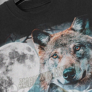 以下が含まれることがあります： 月に向かって吠えるオオカミのグラフィックプリントが施された黒いTシャツ。月は薄い青色で、オオカミは茶色と灰色です。シャツには「SEACOAST」という文字がプリントされています。