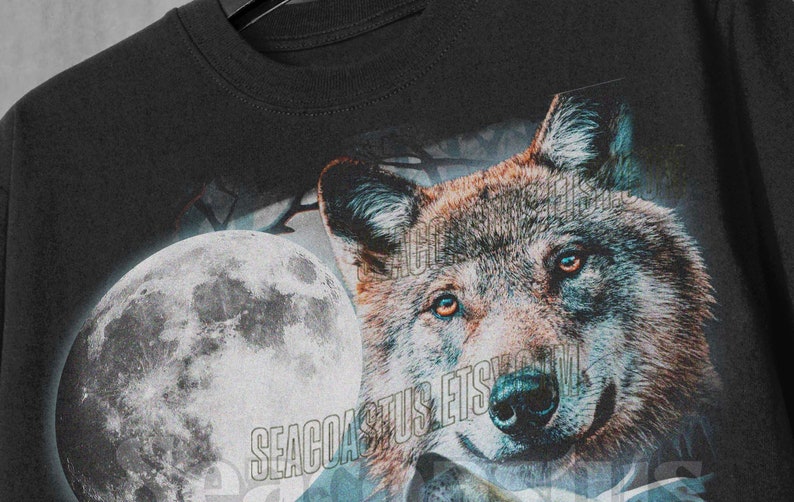 以下が含まれることがあります： 月に向かって吠えるオオカミのグラフィックプリントが施された黒いTシャツ。月は薄い青色で、オオカミは茶色と灰色です。シャツには「SEACOAST」という文字がプリントされています。