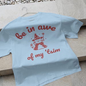 May include: Light blue t-shirt with a red graphic of a cat wearing a witch hat and holding a wand. The text "be in awe of my 'tsim" is printed in red on the shirt.