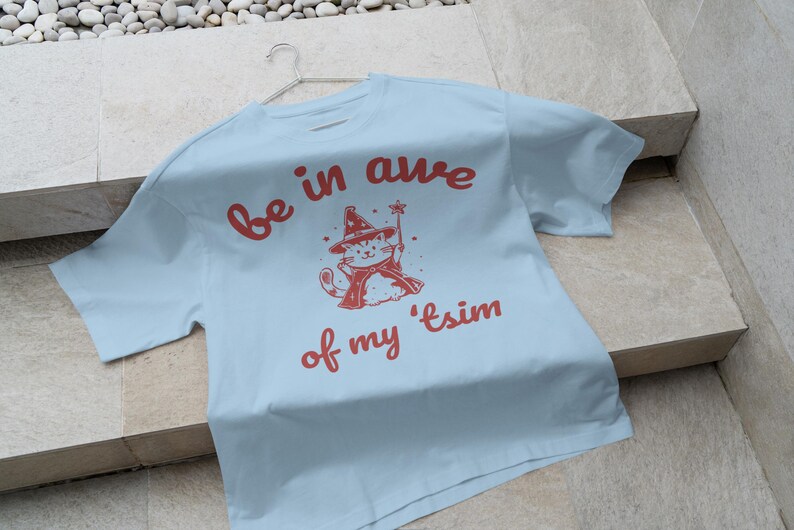 May include: Light blue t-shirt with a red graphic of a cat wearing a witch hat and holding a wand. The text "be in awe of my 'tsim" is printed in red on the shirt.