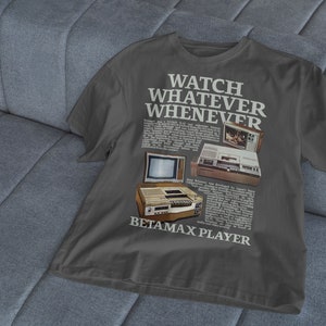 May include: A dark grey t-shirt featuring a vintage advertisement for a Betamax player. The text on the shirt reads "Watch Whatever Whenever Betamax Player".