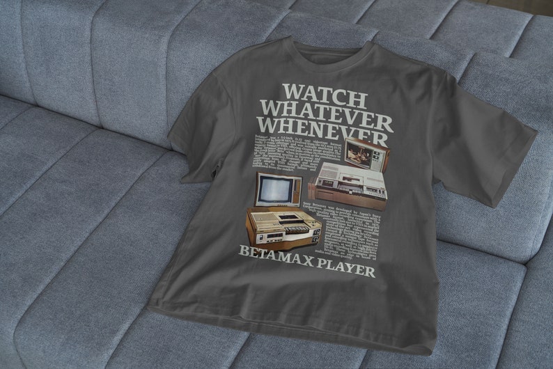 May include: A dark grey t-shirt featuring a vintage advertisement for a Betamax player. The text on the shirt reads "Watch Whatever Whenever Betamax Player".