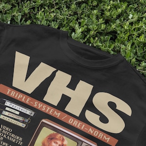 May include: Black t-shirt with a vintage-style graphic of a VHS tape and television set. The text reads "VHS Triple-System / Orei-Norm" and "Preserve your precious memories!"
