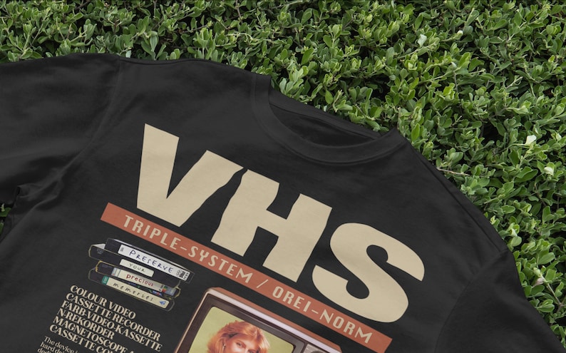 May include: Black t-shirt with a vintage-style graphic of a VHS tape and television set. The text reads "VHS Triple-System / Orei-Norm" and "Preserve your precious memories!"