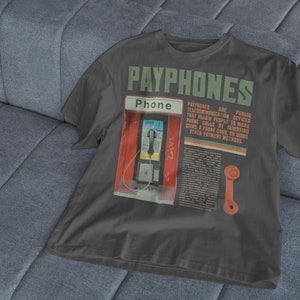 May include: Dark gray t-shirt featuring a graphic of a payphone booth with the word "PAYPHONES" in green. The shirt includes text describing payphones as public telecommunication devices.